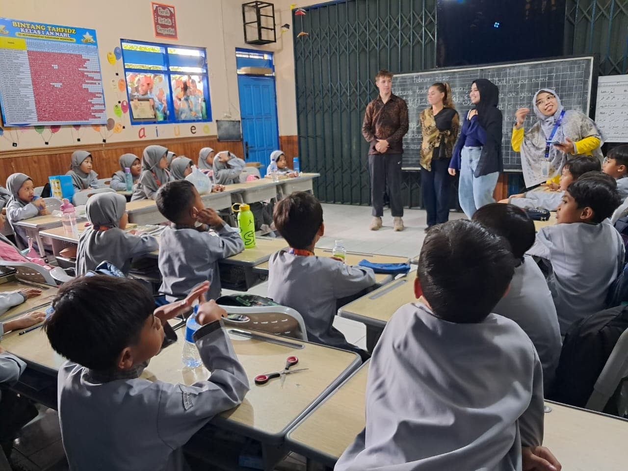 Young students in grey uniforms and hijabs attentively watch five adults in a classroom.