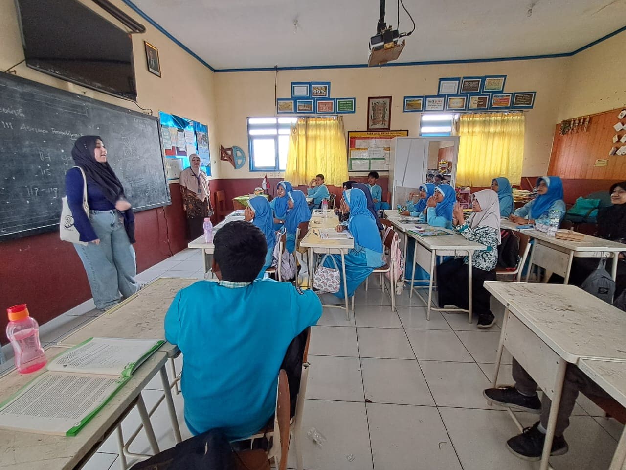 Woman in hijab instructs students in blue uniforms in a bright classroom.